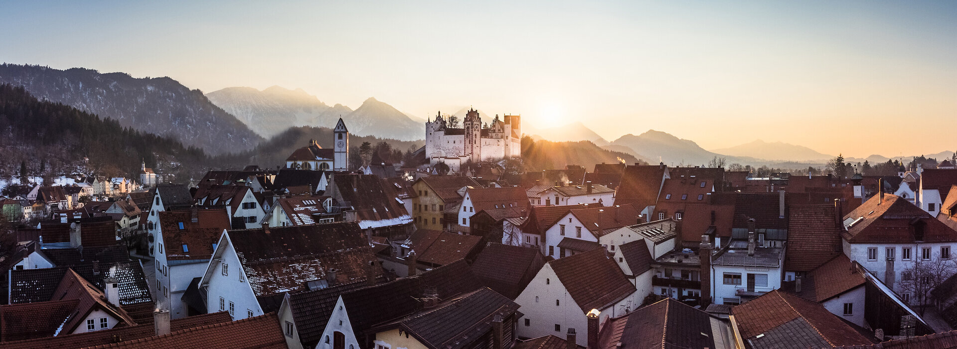 Panoramablick über rote Dächer der Füssener Altstadt. Die winterliche Morgensonne kommt hinter den schneebedeckten Bergen und dem Schloss hervor.