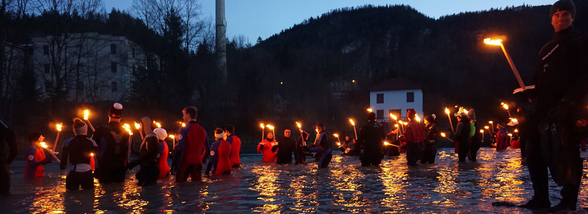 Das Bild zeigt mehrere Personen in Neoprenanzügen im knietiefen Wasser stehen. Alle tragen unterschiedliche Mützen und halten brennende Fackeln in den Händen. Es Dämmert. Es ist schon fast dunkel.  