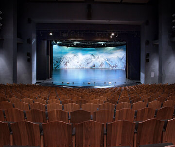 Bühne Festspielhaus Das Foto zeigt den Saal des Festspielhauses Neuschwanstein. Der Blick geht über schier unzählige, braune Holzstuhllehnen hinweg zur Bühne. Das Bühnenbild zeigt schneebedeckte Berge. Der Zuschauerraum ist menschenleer.