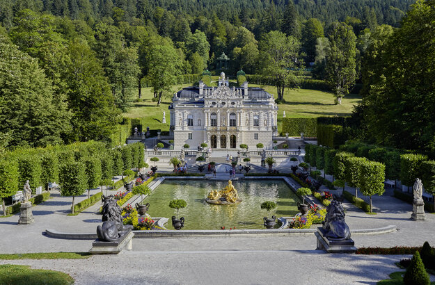 Das Bild zeigt eine Aussenansicht des Schloss Linderhof. Im Vordergrund ist der prächtige Schlossgarten mit einem großen Brunnen zu sehen.