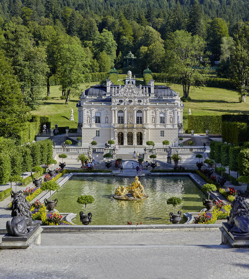 Das Bild zeigt eine Aussenansicht des Schloss Linderhof. Im Vordergrund ist der prächtige Schlossgarten mit einem großen Brunnen zu sehen.