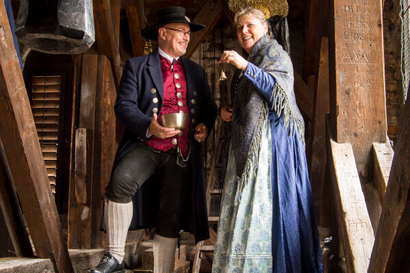 Zwei Personen in traditioneller Kleidung stehen unter einer großen Glocke. Der Mann mit einem Zylinder und einer Schale lächelt die Frau an, die einen eleganten, blauen Rock trägt und ebenfalls lächelt. Die Umgebung ist rustikal mit Holzbalken und einem gewachsten Fußboden.
