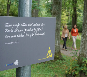 Das Bild zeigt zwei Frauen, die auf einem Waldweg laufen. Ringsherum wachsen hohe Bäume. Auf dem Schild im Vordergrund steht „Man prüfe alles und nehme das Beste. Dieser Grundsatz führt uns am sichersten zur Wahrheit. Sebastian Kneipp“