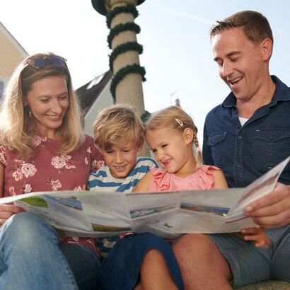  Das Bild ist ein Farbfoto, das eine Familie von vier Personen zeigt, die draußen sitzen. Die Frau, die wahrscheinlich die Mutter ist, ist auf der linken Seite des Bildes zu sehen, sie sitzt und hält eine Zeitung aufgeschlagen vor sich. Sie lächelt und sieht in die Kamera. Neben ihr sitzt ein junges Mädchen, ebenfalls lächelnd und in die Kamera blickend. Das Mädchen trägt ein Rosa-Top. Auf der rechten Seite des Bildes sitzt ein Mann, der ebenfalls die Zeitung aufgeschlagen vor sich hält. Er lächelt und sieht in die Kamera. Der Mann trägt ein Blau-Shirt. Im Hintergrund ist ein gelbes Gebäude mit mehreren Fenstern zu sehen. Der Himmel ist klar und blau, was auf einen sonnigen Tag hindeutet. Es sind keine sichtbaren Texte oder Logos im Bild zu sehen.