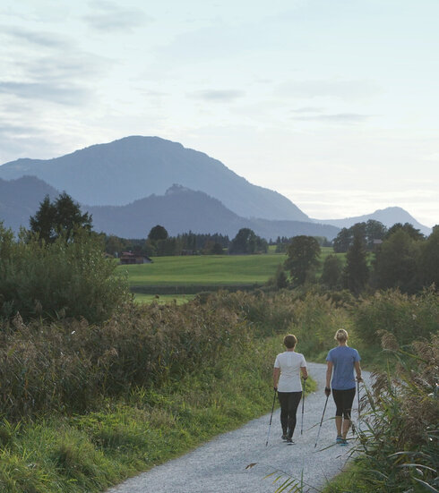 Das Bild zeigt zwei Frauen von hinten die auf einem Kiesweg in einer naturbelassenen Umgebung mit Nordic-Walking-Stöcken laufen. Im Hintergrund ist eine Bergkette zu sehen. 
