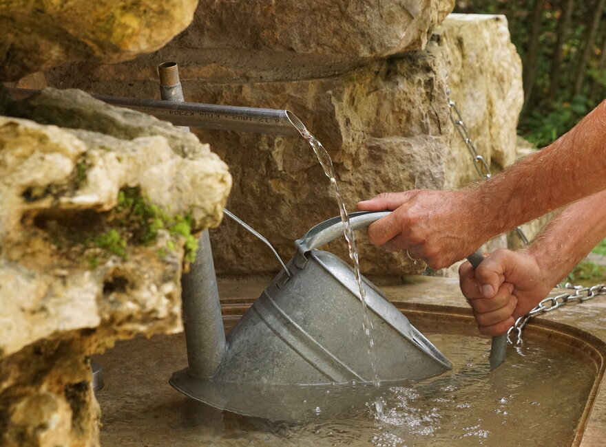 Das Bild zeigt die Nahaufnahme einer Metallgießkanne, die in ein Steinbecken mit Wasser gehalten wird. Aus einem Wasserhahn läuft Wasser herunter in das Becken. Das Becken ist eingerahmt von großen Natursteinen.