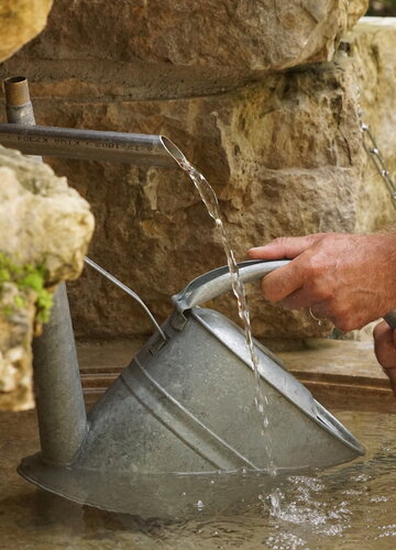 Das Bild zeigt die Nahaufnahme einer Metallgießkanne, die in ein Steinbecken mit Wasser gehalten wird. Aus einem Wasserhahn läuft Wasser herunter in das Becken. Das Becken ist eingerahmt von großen Natursteinen.