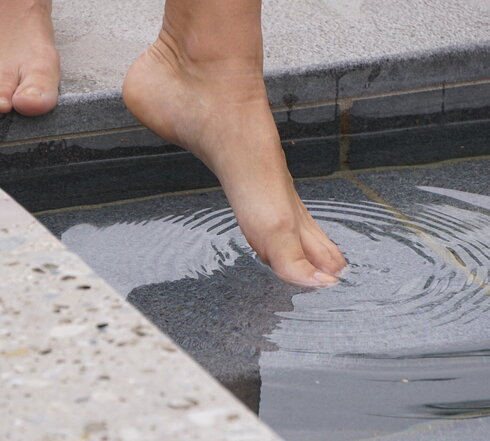 Das Bild eine Person, die vor einem Tretbecken steht und den linken Fuß ins Wasser streckt. Allerdings sind nur die nackten Füße der Person zu sehen. 