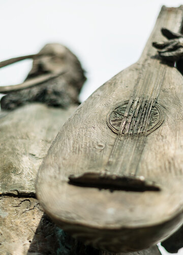 Ein Detail einer Bronzestatue ist zu sehen. Man sieht einen bärtigen Herrn, der eine Laute hält. Die Laute steht im Fokus. Der Rest des Bildes ist unscharf. 