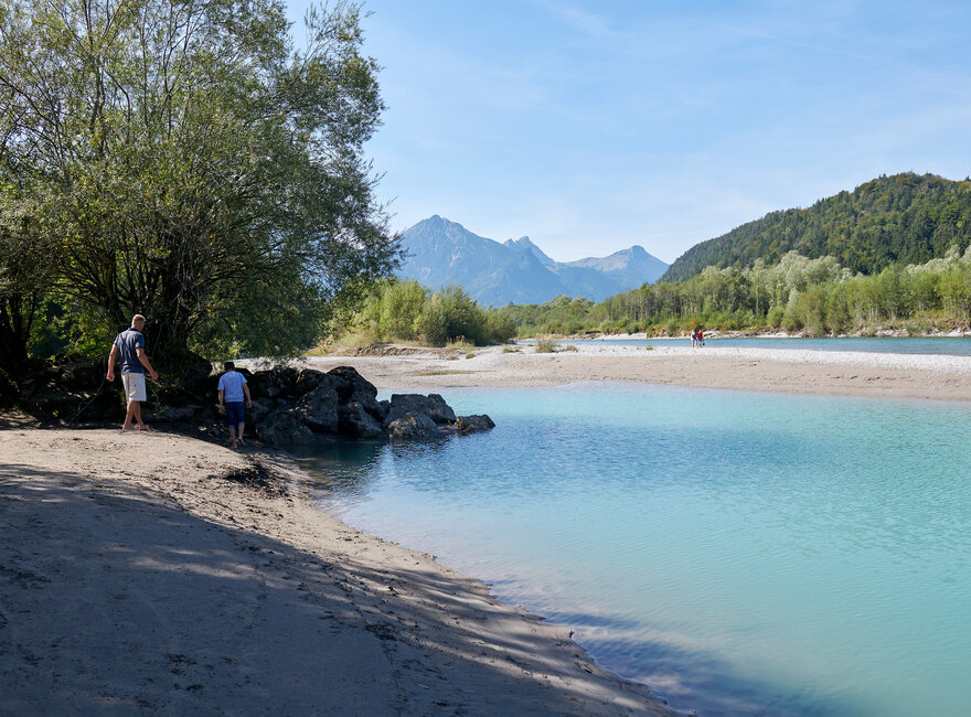 Das Bild zeigt ein Flußbett mit türkisfarbenem Wasser. Am Ufer sind viele Steine. Weiter hinten laufen ein Mann und ein Junge am Ufer entlang.