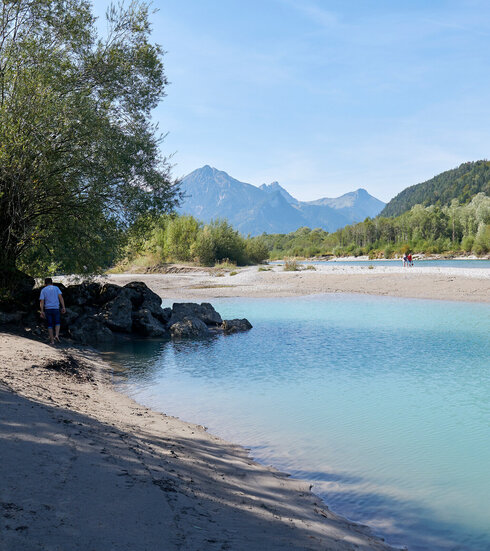 Das Bild zeigt ein Flußbett mit türkisfarbenem Wasser. Am Ufer sind viele Steine. Weiter hinten laufen ein Mann und ein Junge am Ufer entlang.