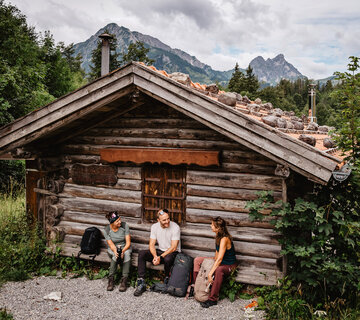 DAs Bild zeigt eine kleine Holzhütte, vor der auf einer Bank zwei Frauen und ein Mann in Outdoor-Kleidung sitzen und sich unterhalten. Im Hintergrund sind Wälder und Berge zu erkennen.