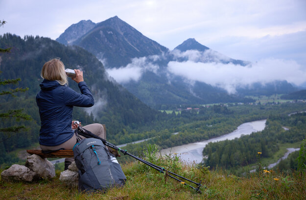 Das Bild zeigt eine Frau, die auf einem Holzscheit auf einer Anhöhe sitzt und ins Tal hinabblickt. Dort fließt ein Fluss. Sie trinkt aus einer Metallflasche und trägt Wanderkleidung. Neben ihr liegen Wanderstöcke. Im Hintergrund erheben sich bewaldete Berge. Der Himmel ist wolkenverhangen. Nebel zieht auf. 