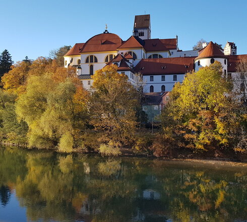 Das Bild zeigt den Blick auf das ehemalige Kloster St. Mang in Füssen im Allgäu vom gegenüberliegenden Lechufer aus. Der brocke Klosterbau erhebt sich über dem herbstlich gefärbten Baumbestand entlang des Flusses Lech. 