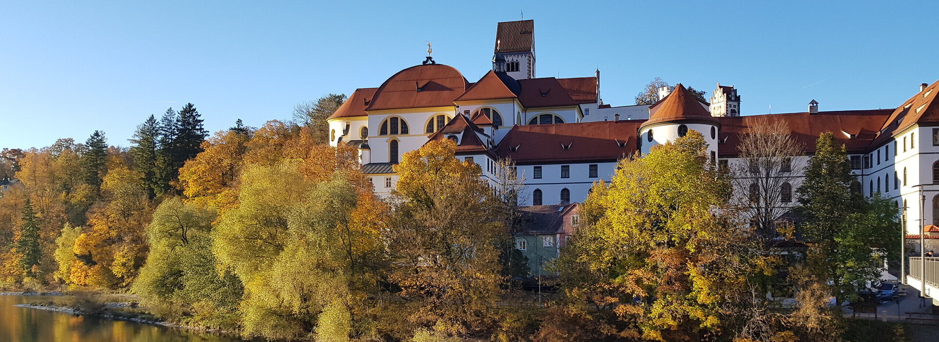 Das Bild zeigt den Blick auf das ehemalige Kloster St. Mang in Füssen im Allgäu vom gegenüberliegenden Lechufer aus. Der brocke Klosterbau erhebt sich über dem herbstlich gefärbten Baumbestand entlang des Flusses Lech. 