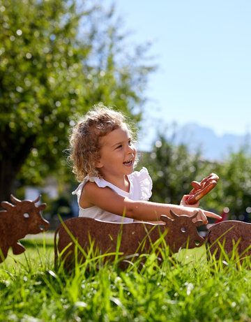 Das Bild zeigt ein Mädchen, das in einer grünen Wiese sitzt und eine Breze in der Hand hat. Im Vordergrund befinden sich 3 kleinere rostige Gartenstecker in Form von Kühen.