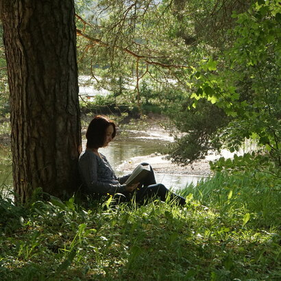 Das Bild zeigt eine Frau, die in einer grünen hohen Wiese sitzt und ein Buch liest. Die Frau lehnt mit dem Rücken an einem großen Baum, der einen weiten Schatten wirft. Zwischen den herabhängenden Ästen kann man im Hintergrund im Sonnenlicht glitzerendes Wasser sehen.