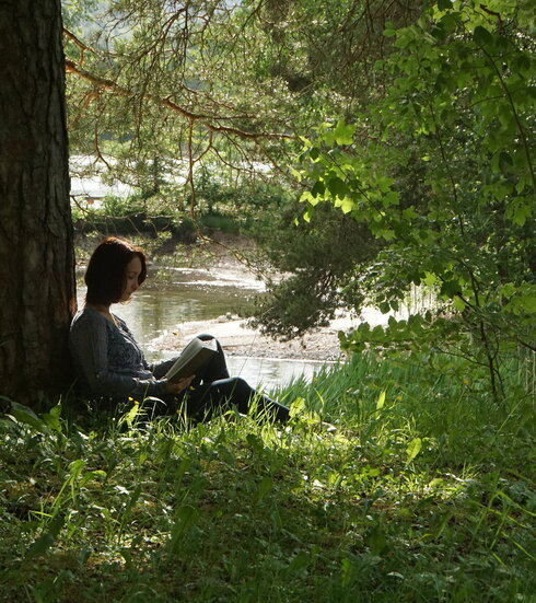 Das Bild zeigt eine Frau, die in einer grünen hohen Wiese sitzt und ein Buch liest. Die Frau lehnt mit dem Rücken an einem großen Baum, der einen weiten Schatten wirft. Zwischen den herabhängenden Ästen kann man im Hintergrund im Sonnenlicht glitzerendes Wasser sehen.