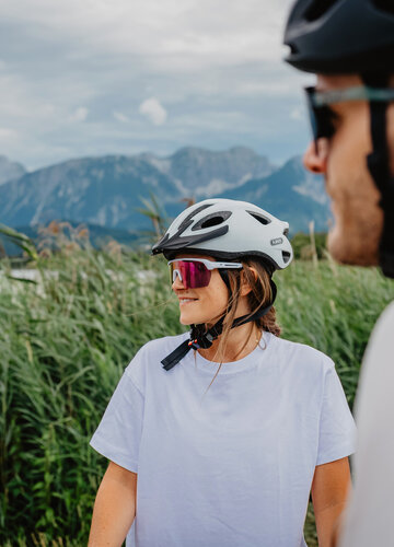 Das Bild zeigt einen Mann und eine Frau mit Fahrradhelm und Sonnebrille. Im Hintergrund ist ein See und die Berge zu erkennen.