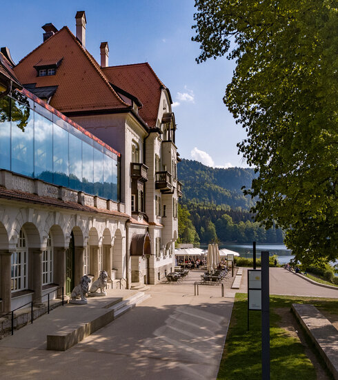 Das Museum der bayerischen Könige am Alpseeufer mit belebter Promenade und Blick auf die Berge.