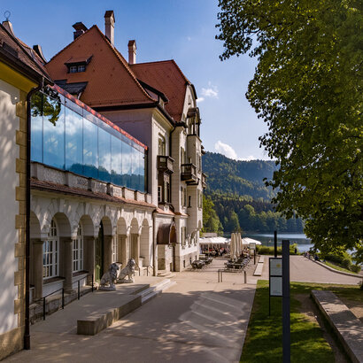 Das Museum der bayerischen Könige am Alpseeufer mit belebter Promenade und Blick auf die Berge.