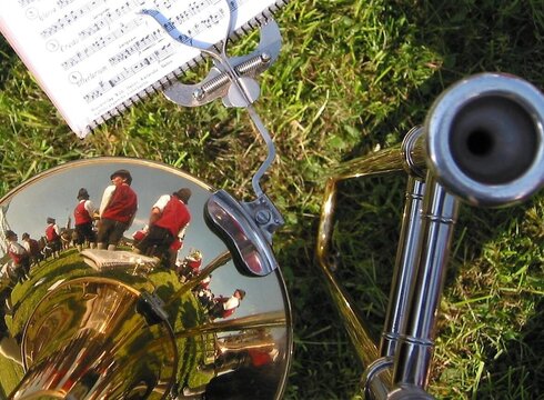 Eine Trompete liegt auf dem Gras, deren Oberfläche das Bild von musizierenden Menschen in roten Jacken reflektiert. Im Hintergrund befindet sich ein Notenblatt, das mit einem Notenständer befestigt ist.
