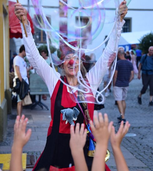 Das Bild zeigt eine Straßenkünstlerin. Sie ist als Clown verkleidet und geschminkt und trägt eine rote Nase. In ihren Händen hält sie zwei Holzstäbe in die Höhe. Vor ihr schweben riesige Seifenblase. Im Vordergrund recken Kinder ihre Hände und Arme in die Höhe. Die Sonne scheint. 