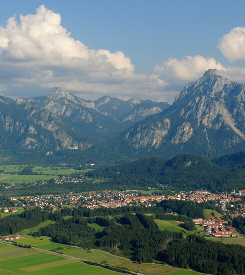 Das Foto zeigt Füssen von oben, umrahmt von Bergen, Hügeln, grünen Wiesen und Wäldern. Auf einer Anhöhe über Füssen thront das Hohe Schloss. Im Hintergrund ist das Schloss Neuschwanstein zu sehen, dass auf einer Erhebung vor den Bergen steht. 