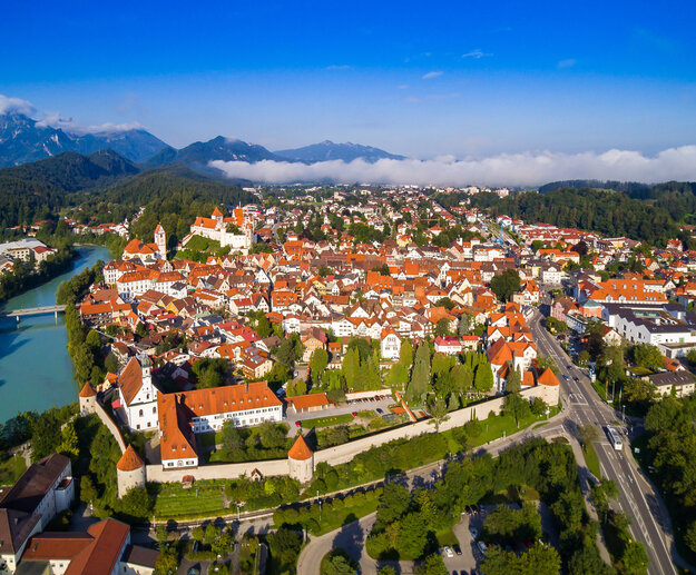 Das Foto zeigt Füssen von oben. Den Blick des Betrachters wird über die Dächer von Füssen geleitet. Im Vordergrund ist das Franziskanerkloster zu erkennen, das aus Klostergebäude und Klosterkirche St. Stephan besteht. Das Klostergelände ist eingerahmt von der historischen Stadtmauer. Neben dem Kloster fließt der türkisblau Fluss Lech. Im Hintergrund des Klosters erstreckt sich die Füssener Altstadt mit dem Hohen Schloss und dem Bergpanorama.