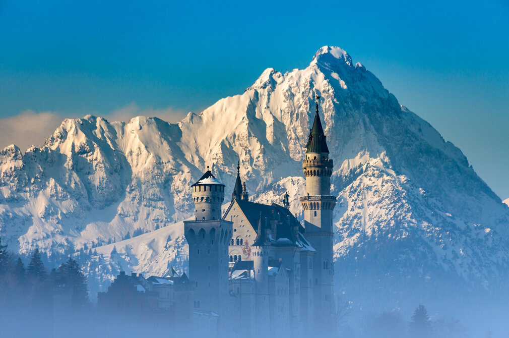 Das weiße Schloss Neuschwanstein mit hohen Türmen und spitzen Dächern vor schneebedeckten Bergen und tiefblauem Himmel.