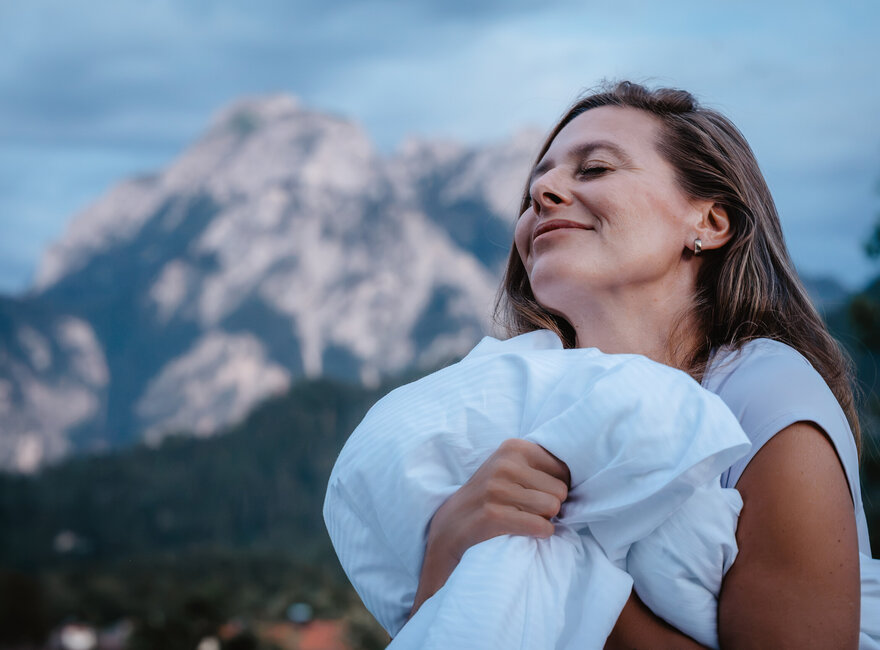 Das Bild zeigt eine Frau mit geschlossenen Augen vor einer Bergkulisse, die eine Bettdecke in den Händen hält.