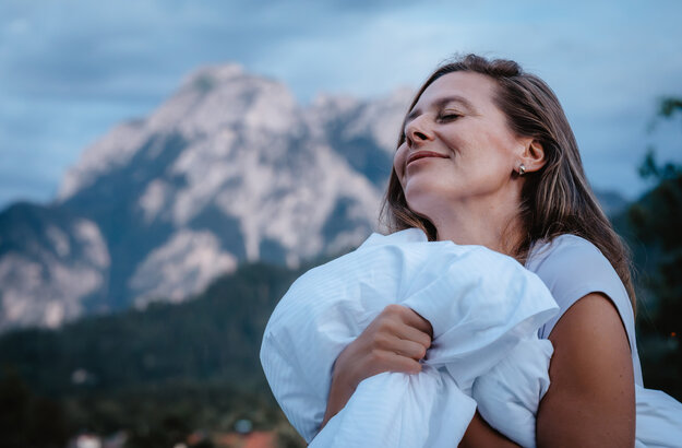 Das Bild zeigt eine Frau, die mit einer Bettdecke in der Hand mit geschlossenen Augen vor einer Bergkulisse steht