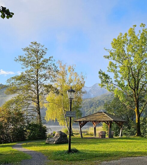 Vom Betrachter führen zwei Kieswege weg. Einer führt nach rechts außerhalb des Blickfelds, der andere geradeaus auf einen hölzernen Pavillon zu, der auf einer Wiese steht. Um den Pavillon herum stehen grüne Laubbäume und Büsche. Im Hintergrund erheben sich bewaldete Hügel und Berge. Der Himmel ist blau. Über dem Wasser des Sees, liegt noch der Morgennebel. Die Sonne scheint.    