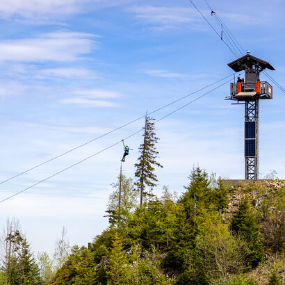 Das Bild zeigt eine Outdoor-Zipline. Man sieht einen großen Turm von dem ein Seil über die Baumwipfel führt. An dem Seil hängt eine Person.