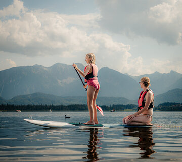 Das Bild zeigt eine Frau und ein Mädchen auf einem SUP mitten auf einem See. Beide tragen Badekleidung und eine Schwimmweste. Das Mädchen steht auf dem SUP und hat ein Paddel in der Hand. Hinter ihr kniet die Frau und blickt in die Ferne. Im Hintergrund erheben sich die Berge. Der Himmel ist blau, aber mit Wolken durchzogen. 