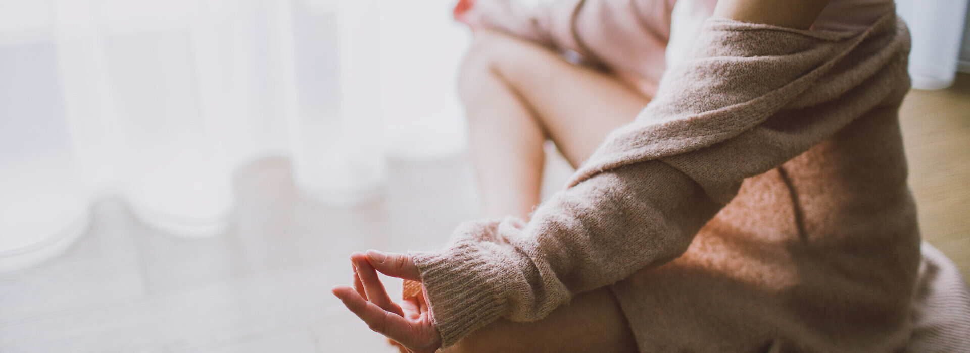 Das Bild zeigt eine Frau, die im Schneidersitz auf dem Holzboden sitzt. Sie sitz vor einem hellen Fenster, das mit einem bodenlangen Stohr verhängt ist. Sie eine beige Strickjacke, die ihr von den Schultern gerutscht ist, ein rosafarbenes Tanktop und eine kurze Hose. Ihre Hände ruhen auf ihren Knien. Mittelfinger und Daumen beider Hände berühren sich.     
