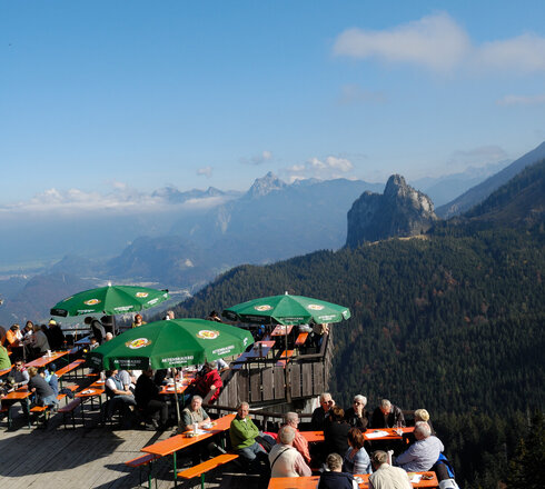 Das Bild zeigt eine Holzterrasse hoch oben auf einem Berg. auf der Terrasse sitzen Menschen auf Bierbänken und Tischen unter grünen Sonnenschirmen.