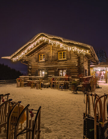 Rodel stehen im Schnee vor der mit Lichterketten am Dach warm erleuchteten Berghütte "Drehhütte". 