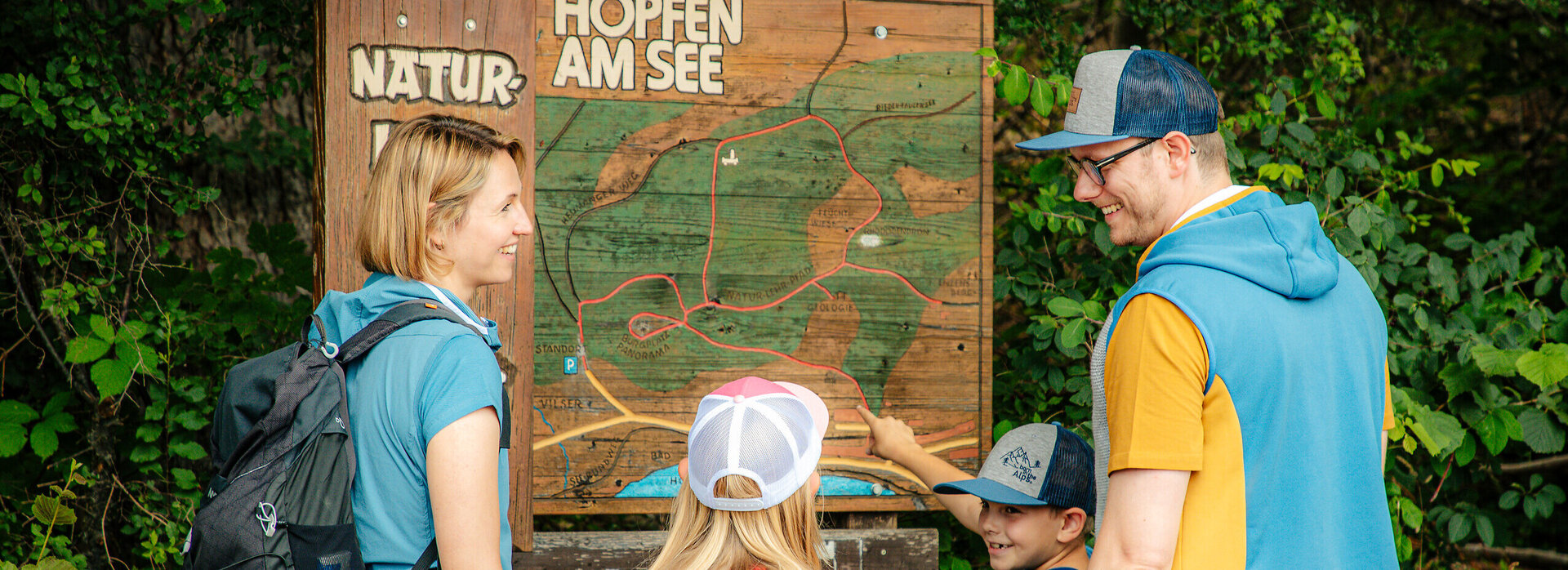 Das Bild zeigt eine Familie vor einer Holztafel mit Informationen zum Hopfensee. Sie tragen Wanderbekleidunge 