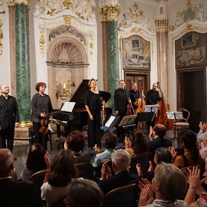 Das Bild zeigt eine Gruppe Musiker auf einer niedrigen Bühne stehen. Einige haben Geigen in den Händen. Auch ein Cello und ein Kontrabass ist dabei. Im Hintergrund steht ein Konzertflügel.  Die Wände des Raums sind prunkvoll verziert. Vor der Bühne sitzen zahlreiche Personen auf Stühlen der Bühne zugewandt und applaudieren. Der Raum ist hell erleuchtet. 