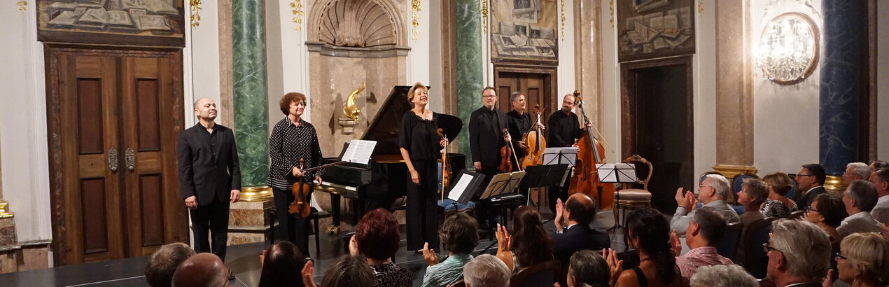Das Bild zeigt eine Gruppe Musiker auf einer niedrigen Bühne stehen. Einige haben Geigen in den Händen. Auch ein Cello und ein Kontrabass ist dabei. Im Hintergrund steht ein Konzertflügel.  Die Wände des Raums sind prunkvoll verziert. Vor der Bühne sitzen zahlreiche Personen auf Stühlen der Bühne zugewandt und applaudieren. Der Raum ist hell erleuchtet. 