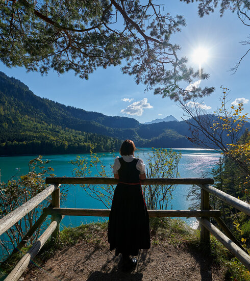 Eine Frau steht an einem Aussichtspunkt am Ufer des türkisgrünen Lechs in Füssen
