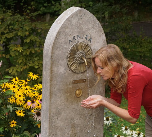 Man blickt auf eine Frau, die sich etwas bückt, um mit ihren Händen, die zu einer Schale geformt sind, das Wasser eines Brunnens aufzufangen. Der Brunnen besteht aus einer Spitz zulaufenden Steinplatte. Im oberen Drittel ist eine bronzene Arnikablüte eingelassen, aus deren Mitte ein kleiner Wasserhahn ragt. Im Hintergrund sind blühende Blumen und Bäume zu sehen. 