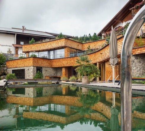 Das Bild zeigt den luxuriösen Außenbereich eines Hotels mit einem Schwimmbad. Das Becken ist der zentrale Fokus des Bildes und wird umgeben von einer Holzterrasse. Die Terrasse ist mit mehreren Töpfen bepflanzt, was einen grünen Akzent in die Szene bringt. Auf der rechten Seite des Bildes ist eine Holzbank zu sehen, die einen Ort zum Entspannen bietet. Das Schwimmbadgebiet wird noch weiter durch eine Holzlaube aufgewertet, die Schatten spendet und zur Gesamtausstattung des Raumes beiträgt. Der Hintergrund des Bildes zeigt ein großes Haus, dem Biohotel Eggensberger in Hopfen. Das Haus, mit seiner Holzfassade, ergänzt das Schwimmbad, wodurch eine harmonische Verbindung von Architektur und Natur entsteht.