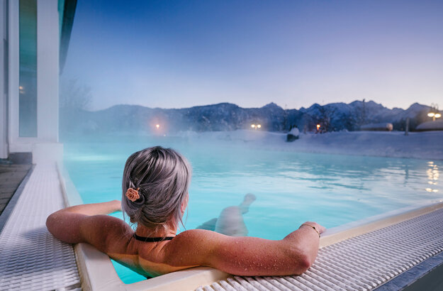 Das Bild zeigt eine Frau, die in Badekleidung in einem dampfenden Outdoor-Pool liegt. Die Landschaft im Hintergrund ist tief verschneit.  