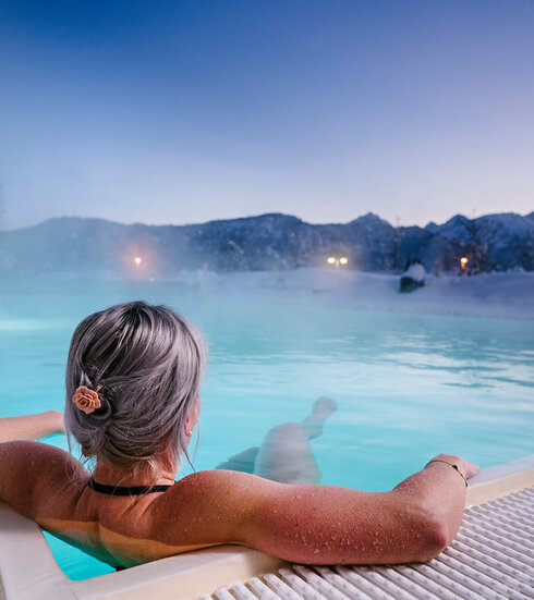Das Bild zeigt eine Frau, die in Badekleidung in einem dampfenden Outdoor-Pool liegt. Sie dreht dem Betrachter den Rücken zu und hat die Haare hochgesteckt. Ihre Arme ruhen auf dem Poolrand. Die Landschaft im Hintergrund ist tief verschneit.  