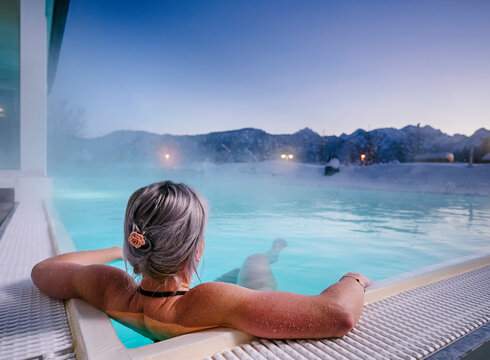 Das Bild zeigt eine Frau, die in Badekleidung in einem dampfenden Outdoor-Pool liegt. Die Landschaft im Hintergrund ist tief verschneit.  