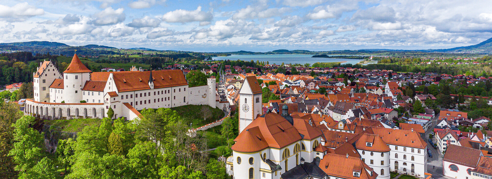 Die Abbildung zeigt eine malerische Stadt mit einer Burgenstruktur, dem Hohen Schloss und auf das alte Kloster St. Mang, die von reichlich grünen Bäumen umgeben ist. Die Stadt ist in einem Tal gelegen, mit einem Fluss, der durch sie hindurchfließt. Im Hintergrund sieht man den Forggensee.