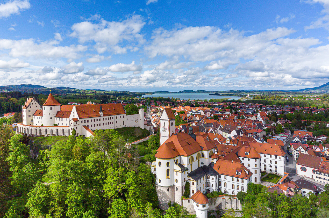 Die Abbildung zeigt eine malerische Stadt mit einer Burgenstruktur, dem Hohen Schloss und auf das alte Kloster St. Mang, die von reichlich grünen Bäumen umgeben ist. Die Stadt ist in einem Tal gelegen, mit einem Fluss, der durch sie hindurchfließt. Im Hintergrund sieht man den Forggensee.