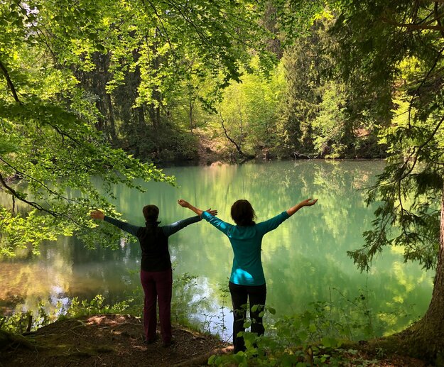 Auf dem Bild sind zwei Frauen, die ihre Arme in die Luft strecken, in einem Wald vor dem Ufer eines Weihers zu sehen